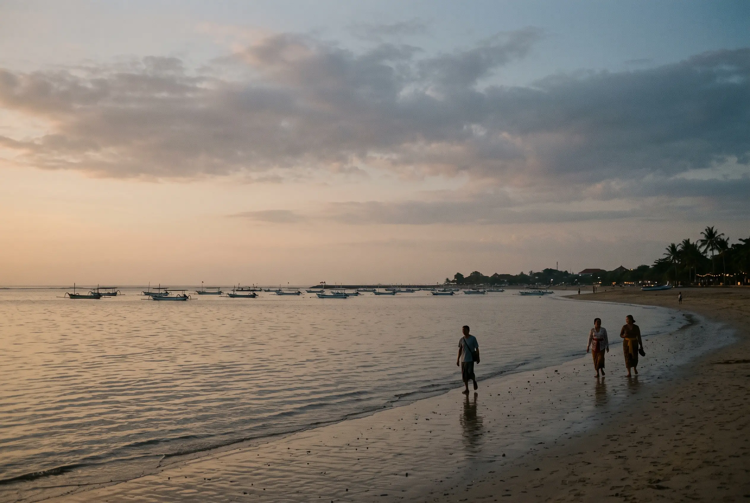 Paseantes junto a barcas de pesca tradicionales en la playa de Sanur al anochecer