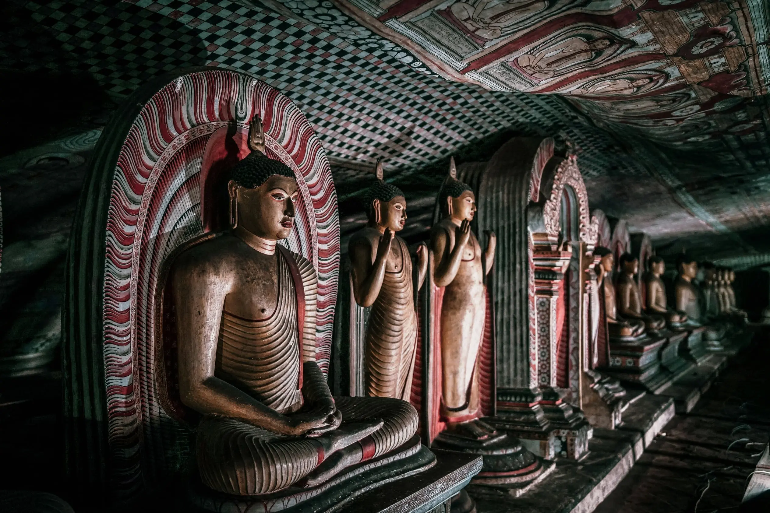 Hilera de estatuas de Buda, sentadas y de pie, bajo el techo de roca pintado de una de las cuevas del templo de Dambulla